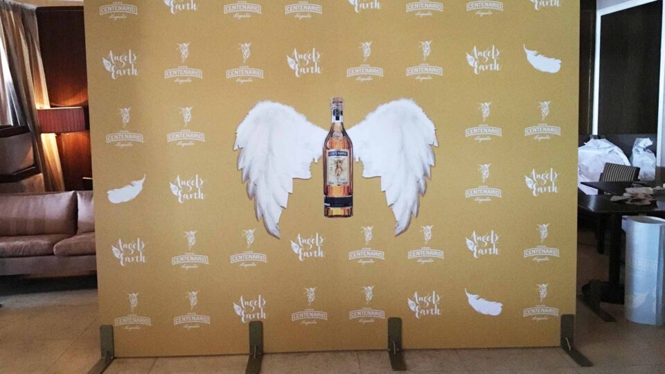 Gold media wall with white Centenario Tequila logo and bottle with wings