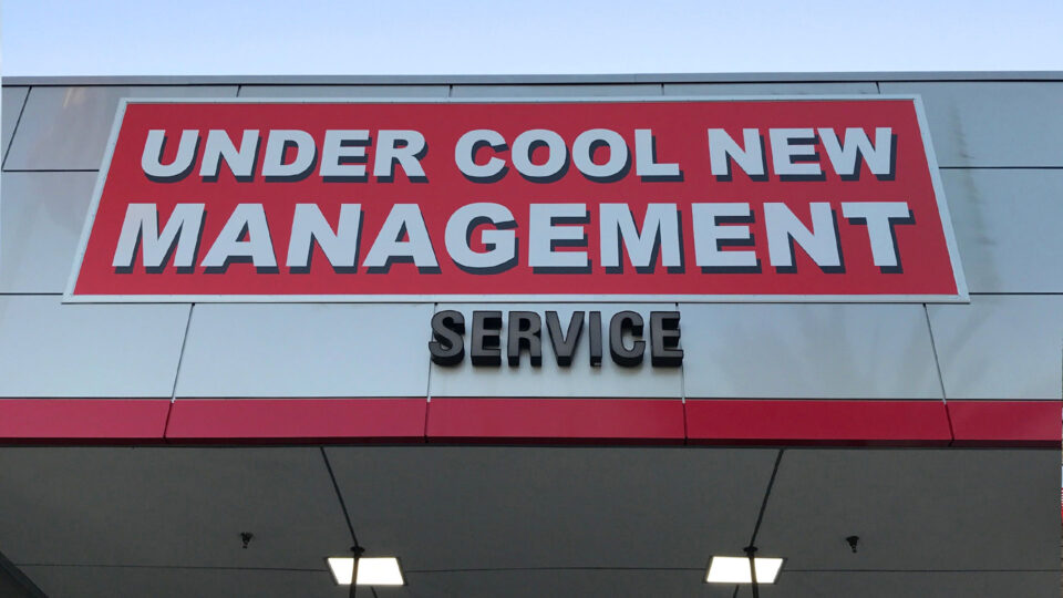 Red and white banner that says Under Cool New Management on top of outside of service department at Toyota dealer.