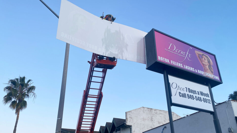 Man on lift changing out acrylic sign. Sign has purple background with model for Derm FX promoting Botox, Fillers, etc.