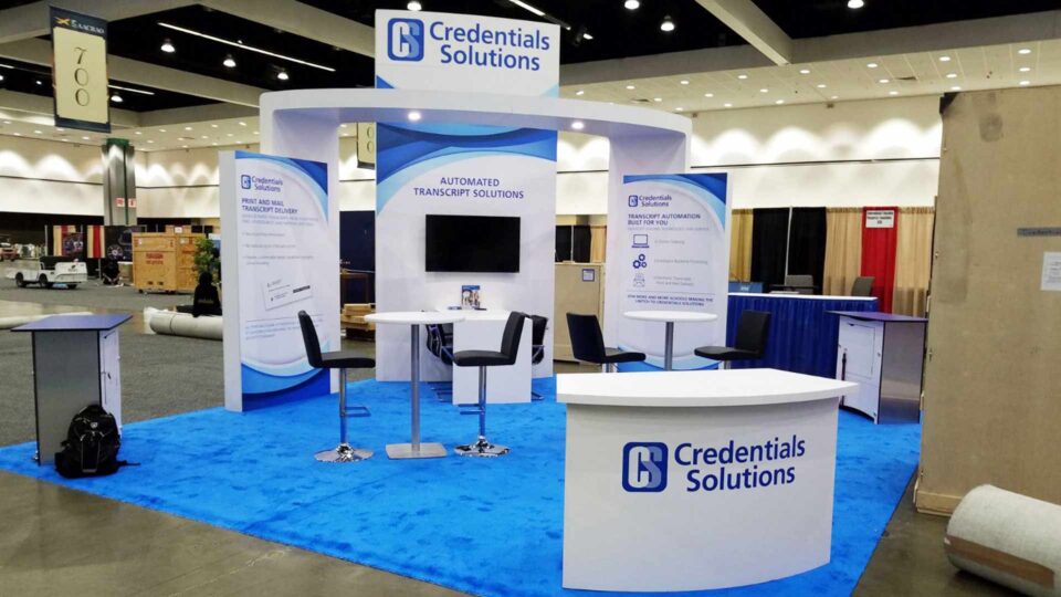 White and Blue trade show booth exhibit with blue carpet and white walls and counter with Credentials Solutions blue vinyl graphics