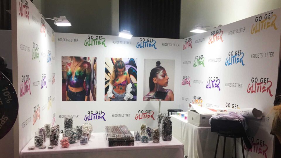 White trade show booth walls with Go Get Glitter logo in mulit colors, along with 3 images of females with colorful body glitter.