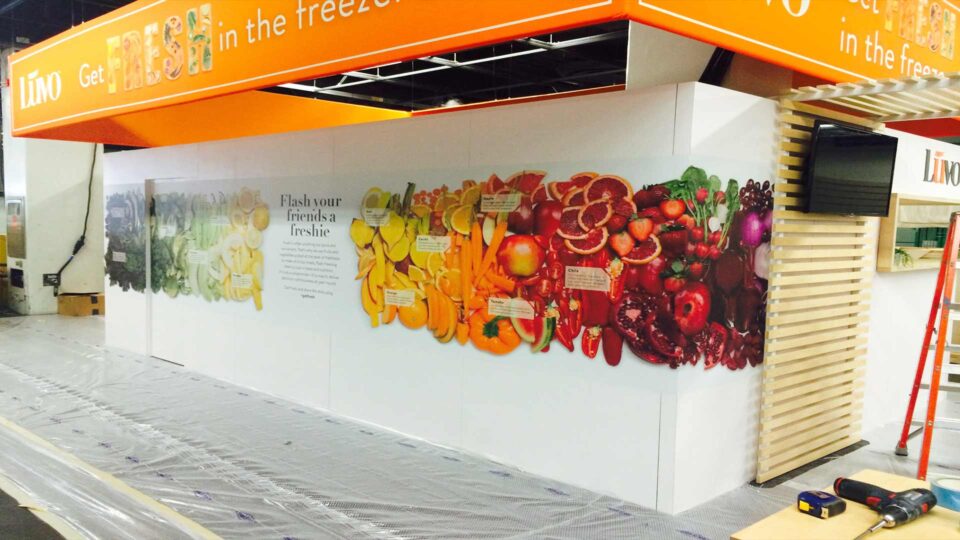 White trade show booth walls with fresh fruits and vegetables. Orange booth topper with Luvo info.
