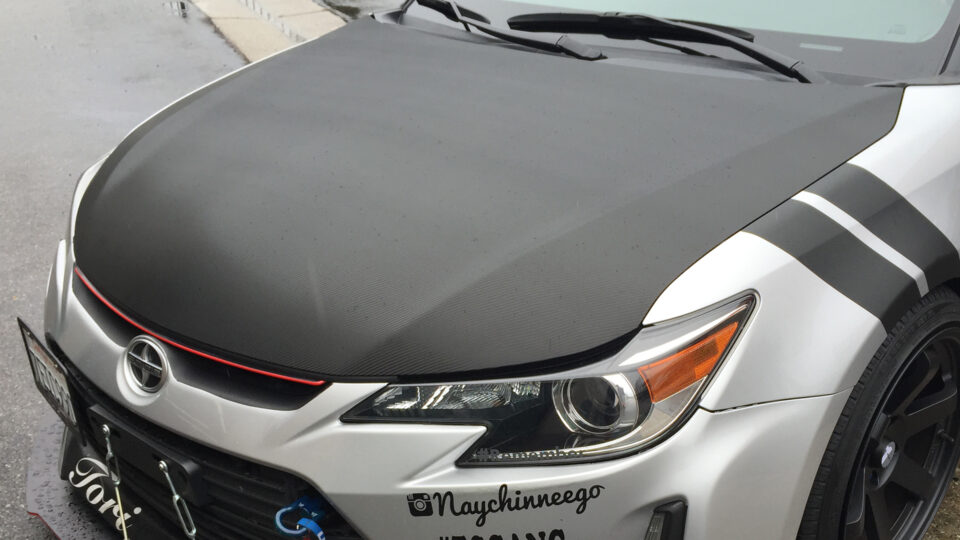 Gray carbon fiber Scion vinyl hood wrap on white car with logos and black stripe