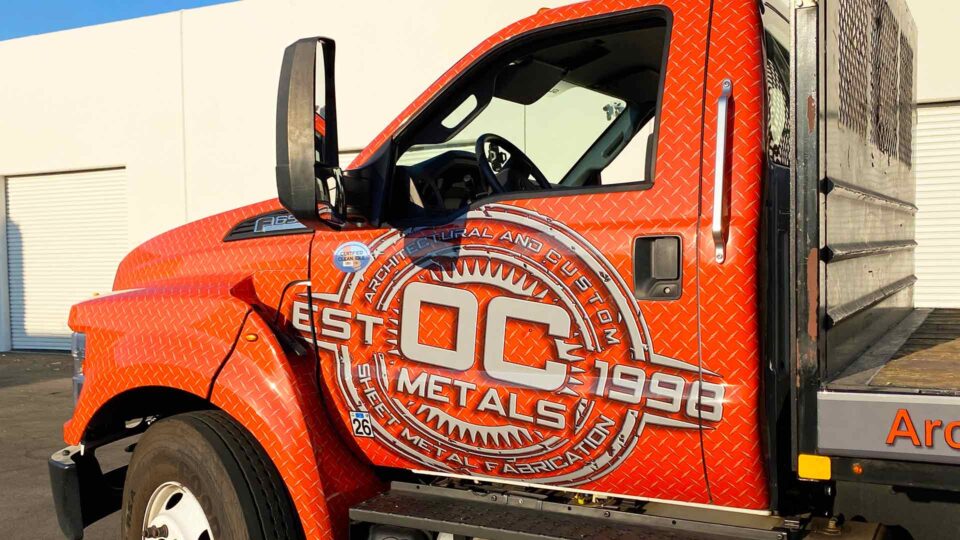 Orange Ford F650 Cab with OC metals logo in white. Services listed on side of flatbed