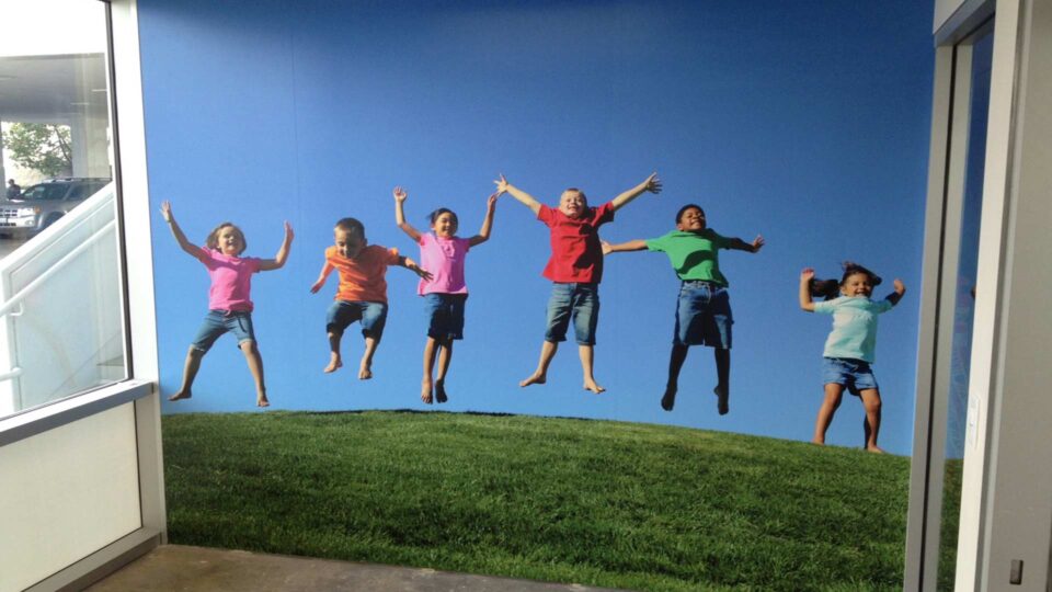 wall graphic of 6 kids in colorful tshirts jumping with hands up over grass with blue sky