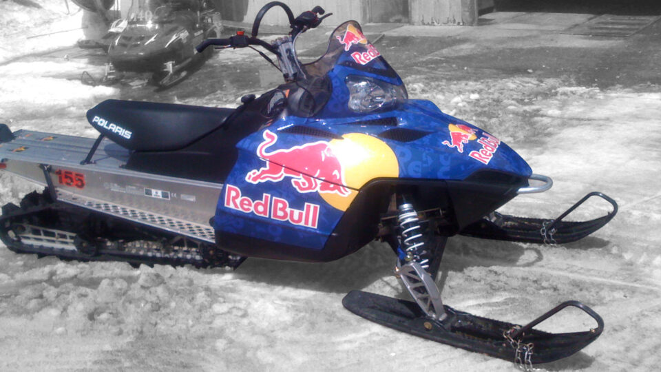 Polaris snowmobile vinyl wrap in blue with Red and yellow red bull logo. Sititng in snow at ski resort.
