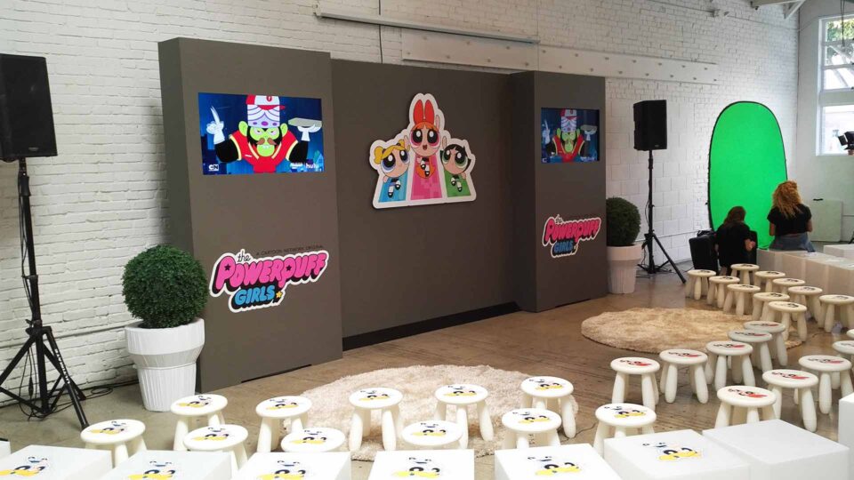 Gray gatorboard TV Wall with Powerpuff girls and logos in room with small stools and speaker for event