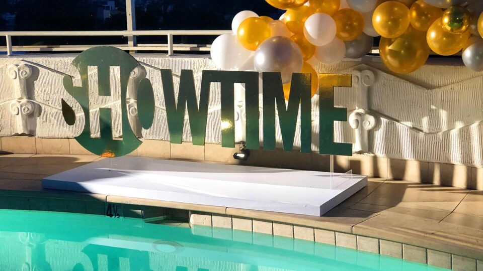 Green router cut acrylic Showtime sign by pool with gold, silver and white balloons for special event