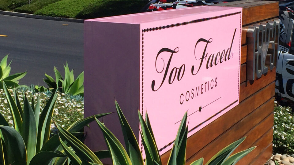 Pink square sign with black text that says Too Faced Cosmetics on wood background with address.