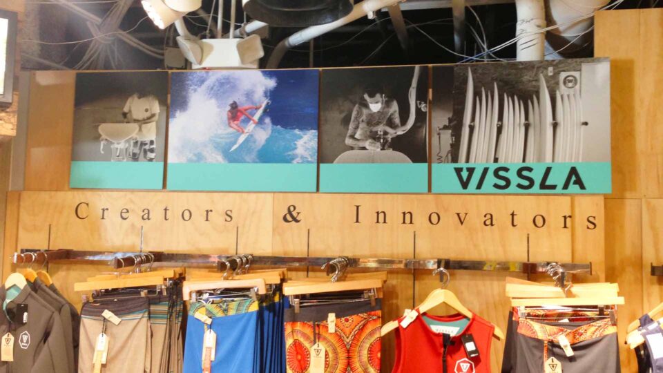 wood wall with printed images of surfing and surfboard manufacturing for VISSLA with mens surf trunks displayed beneath