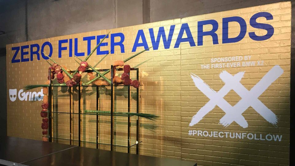 Gold wall with ZERO FILTER AWARDS in blue text with white graphics. Black shelves with pinecones and sharp greenery in front