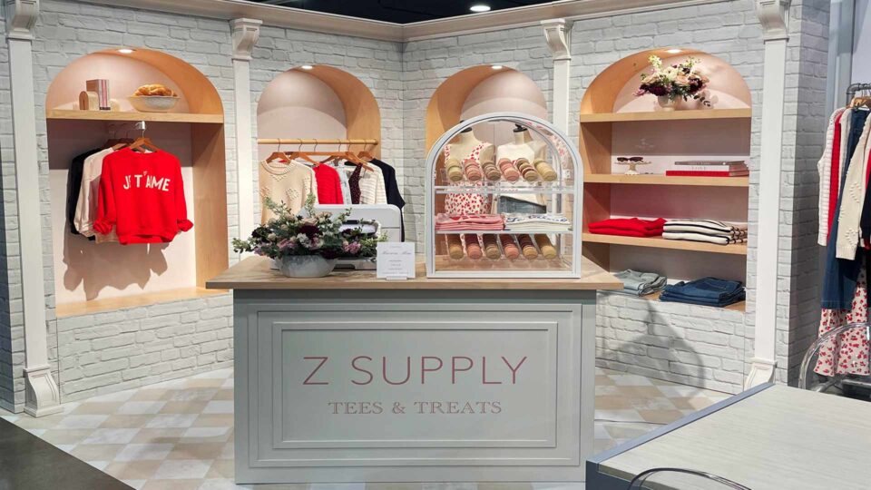 Trade show booth with faux brick vinyl wrap, clothes hanging and folded on shelves. Front desk with Zsupply logo and flowers and cookies