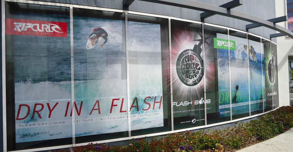 Wall graphics of surfer on waves and Rip Curl Flashbomb Wetsuit logo, inside of surf shop window
